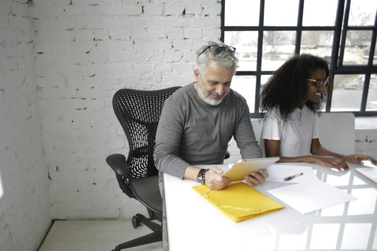 Smiling mature male executive surfing tablet while sitting near African American female colleague in eyeglasses working together in creative office